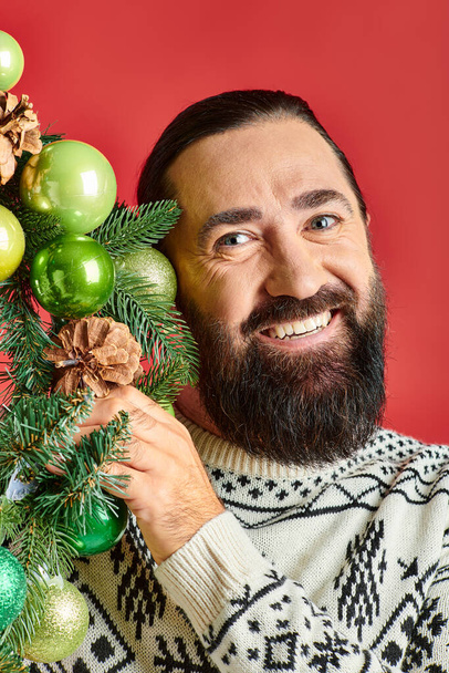 joyful bearded man in winter sweater holding decorated Christmas wreath with baubles on red backdrop - Photo, Image