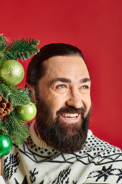 joyful bearded man in Christmas sweater holding decorated wreath with baubles on red backdrop - Photo, Image