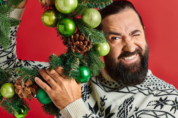 クリスマスセーターで幸せなひげを被った男 赤い背景に問題を抱えた装飾的な羽根を保持 - 写真・画像