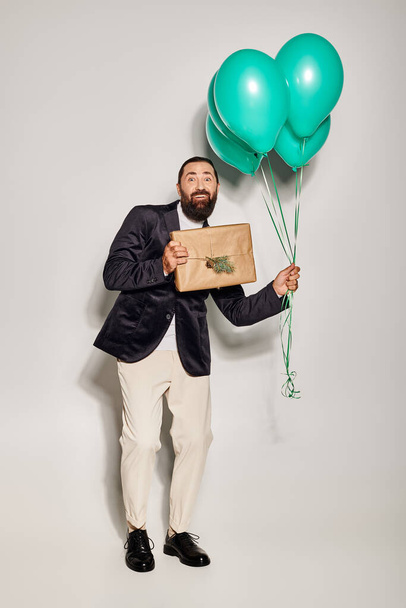 happy bearded man in formal wear holding blue balloons and Christmas  present on grey backdrop - Photo, Image