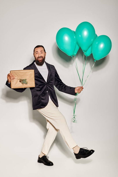 excited bearded man in formal wear holding blue balloons and Christmas  present on grey backdrop - Photo, image