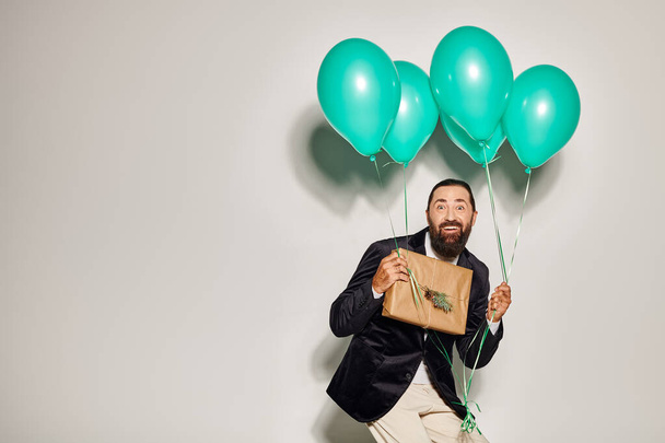 cheerful bearded man in formal wear holding blue balloons and Christmas  present on grey backdrop - Photo, Image