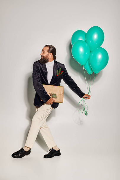 happy bearded man in formal wear holding blue balloons and walking with Christmas present on grey - Photo, Image