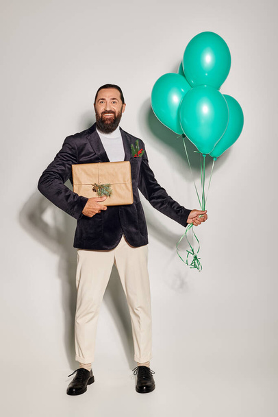 hombre barbudo feliz en ropa formal sosteniendo globos azules y de pie con regalo de Navidad en gris - Foto, imagen