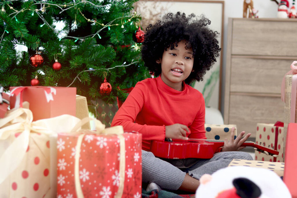 Cute African girl child with curly hair playing ukulele during sitting on floor around many present gift boxes under decorative Christmas tree. Happy kid celebrating winter holiday. Merry Christmas - Photo, Image