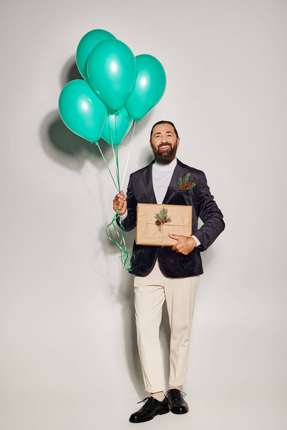 smiling bearded man in formal attire holding blue balloons and Christmas  present on grey backdrop - 写真・画像