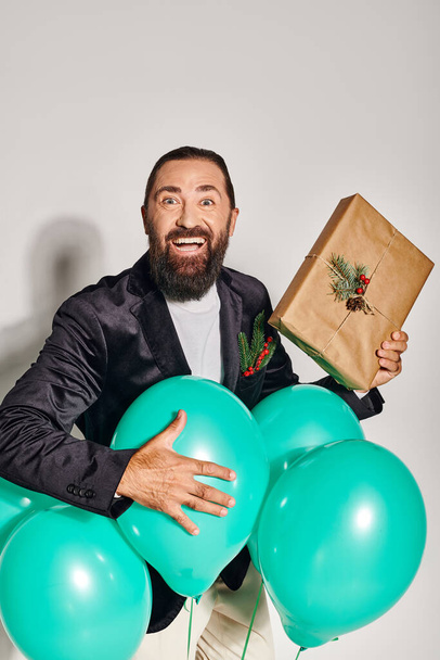 Resmi kıyafetli neşeli sakallı adam, gri arka planda mavi balonların yanında Noel hediyesini tutuyor. - Fotoğraf, Görsel