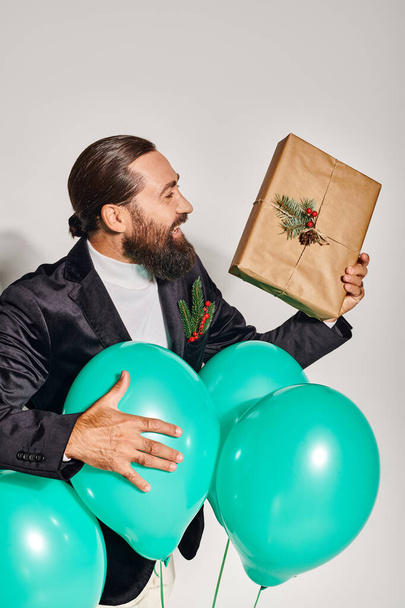 uśmiechnięty brodaty mężczyzna w formalnym ubraniu trzymający prezent świąteczny w pobliżu niebieskich balonów na szarym tle - Zdjęcie, obraz