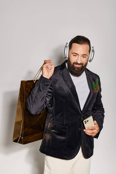 veselý vousatý muž v bezdrátových sluchátkách držící smartphone a nákupní tašku na šedém pozadí - Fotografie, Obrázek