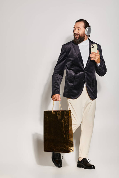 hombre barbudo sonriente en auriculares inalámbricos que sostiene el teléfono inteligente y la bolsa de compras en el fondo gris - Foto, Imagen