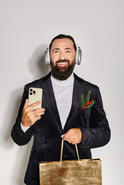 χαρούμενα γενειοφόρος άνθρωπος σε ασύρματα ακουστικά κρατώντας smartphone και τσάντα δώρο σε γκρι φόντο - Φωτογραφία, εικόνα