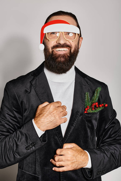 gelukkig baard man in feestelijke glazen met santa hoed poseren in pak en glimlachen op grijze achtergrond - Foto, afbeelding