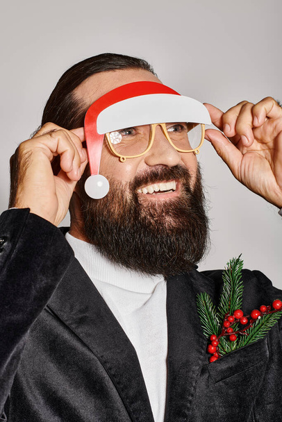 happy bearded man in festive glasses with santa hat posing in suit and smiling on grey backdrop - Photo, Image