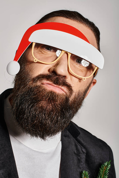 szakállas férfi ünnepi szemüvegben, télapó kalapban, öltönyben pózol, és szürke háttéren duzzog az ajka. - Fotó, kép