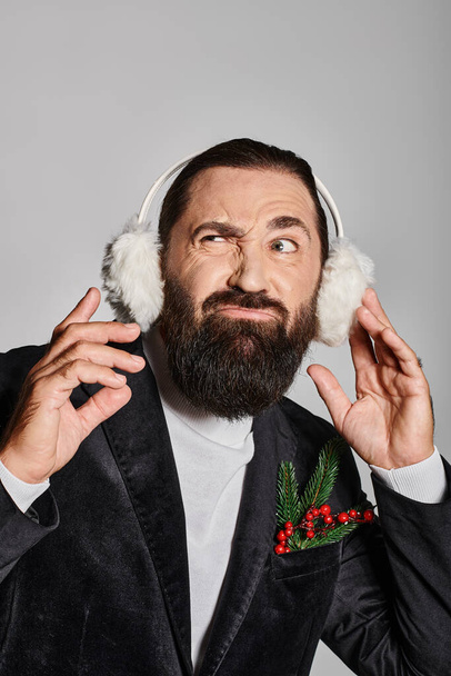 emotional bearded man in suit with Christmas spruce in pocket wearing ear muffs on grey backdrop - Photo, Image