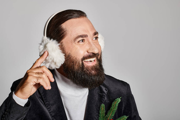 cheerful bearded man in suit with Christmas spruce branch and red berries wearing ear muffs on grey - Photo, Image