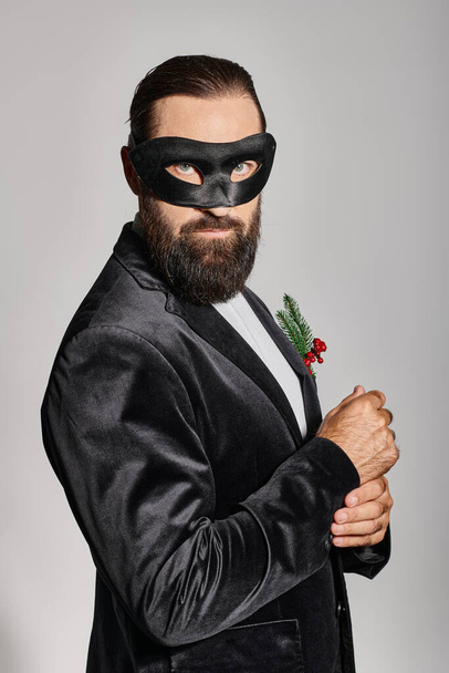Boże Narodzenie bal maskowy, przystojny brodaty mężczyzna w masce karnawałowej i elegancki garnitur na szarym tle - Zdjęcie, obraz