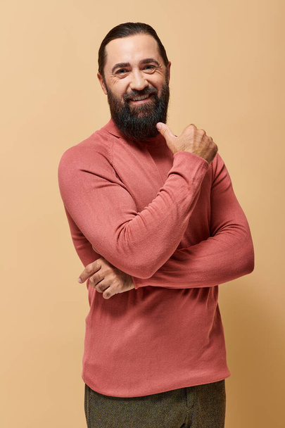 cheerful bearded man in casual turtleneck jumper looking at camera on beige backdrop, male fashion - Photo, Image