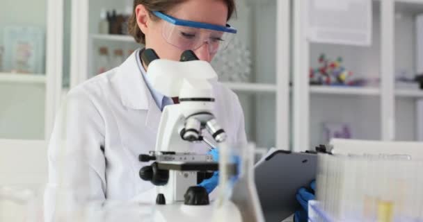 Biological scientist laboratory assistant looks through microscope and writes medical report to laboratory. Chemical and medical research - Footage, Video