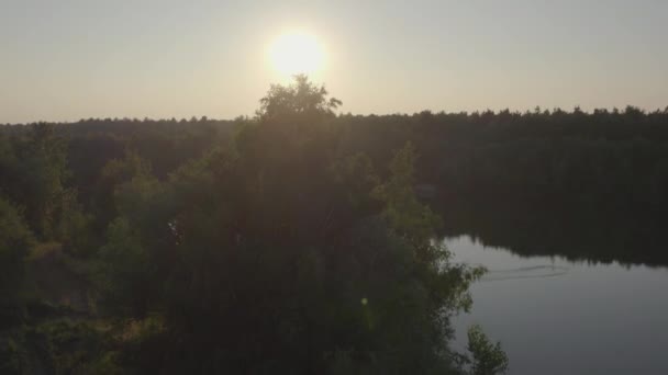 En un suave ascenso, la vista aérea se transforma desde el ocultamiento detrás de un árbol solitario para revelar una impresionante puesta de sol sobre un sereno lago forestal. El cielo de la tarde, pintado con trazos de carmesí - Metraje, vídeo