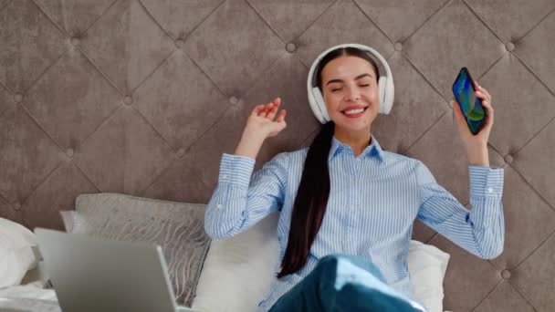 Amazing girl sits on a bed in a hotel, holding a mobile phone, dancing and listening to music. Happy woman selects favorite track from playlist and joyfully spends time dancing along in headphones. - Footage, Video