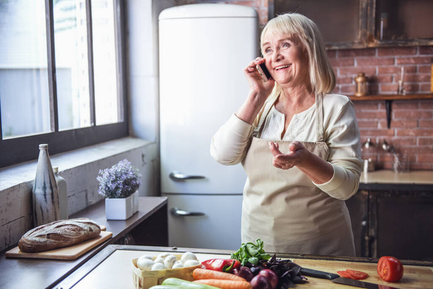 Mooie senior vrouw in schort is praten over de mobiele telefoon en glimlachen tijdens het koken in de keuken - Foto, afbeelding