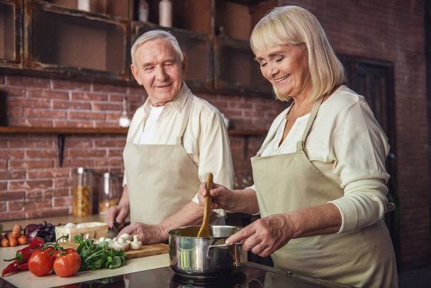 Красива старша пара в фартухах говорить і посміхається, готуючи разом на кухні
 - Фото, зображення