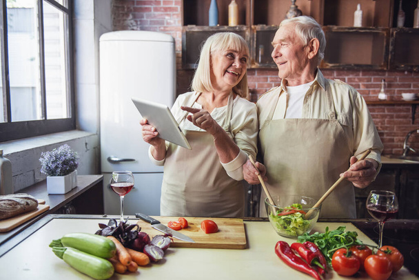 Старшая пара в апронах пользуется цифровым планшетом, разговаривает и улыбается во время совместного приготовления пищи на кухне
 - Фото, изображение