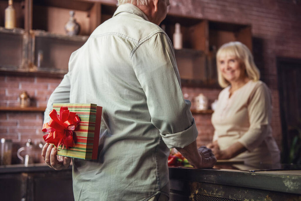 Mujer mayor hermosa está cocinando y sonriendo mientras su marido está sosteniendo un regalo para ella a sus espaldas
 - Foto, imagen