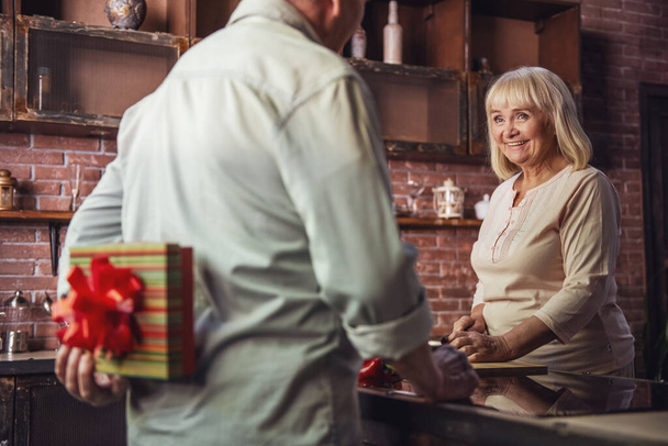 Mulher sênior bonita está cozinhando e sorrindo enquanto seu marido está segurando um presente para ela nas costas
 - Foto, Imagem