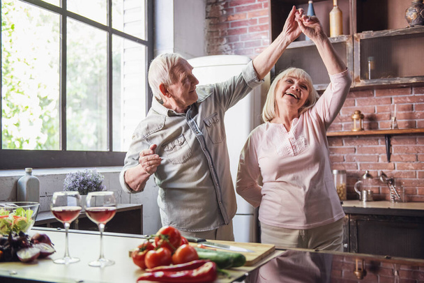 Όμορφο ζευγάρι τελειόφοιτων χορεύει και χαμογελά ενώ μαγειρεύουν μαζί στην κουζίνα - Φωτογραφία, εικόνα