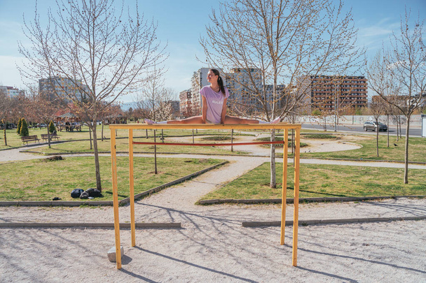 Гибкая спортивная девушка делает упражнения на горизонтальных брусьях в парке - Фото, изображение