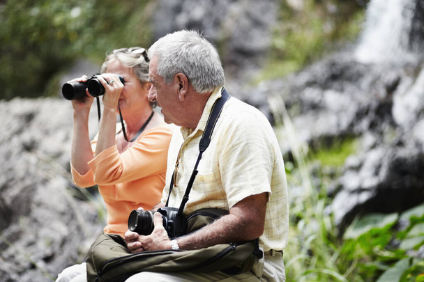 Senior, couple and binocular in forest for tourist, sightseeing or vacation with waterfall and scenery. Elderly, man and woman in woods or nature for holiday, adventure or experience with backpack. - Photo, Image