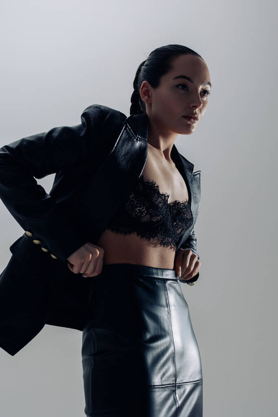 Young pretty woman in black lacy bra, leather skirt and jacket posing against gray background. - Photo, Image