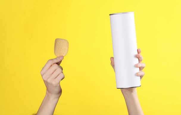 La mano de la mujer sostiene una lata cilíndrica para chips con una etiqueta blanca vacía sobre fondo amarillo. Plantilla para diseño - Foto, imagen