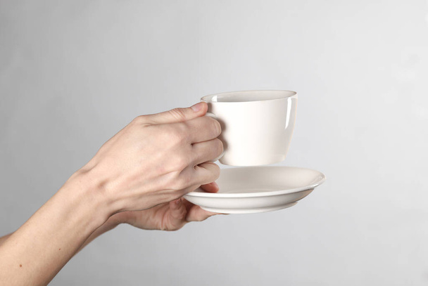 Les mains féminines tiennent une tasse en céramique blanche avec une soucoupe sur fond gris - Photo, image