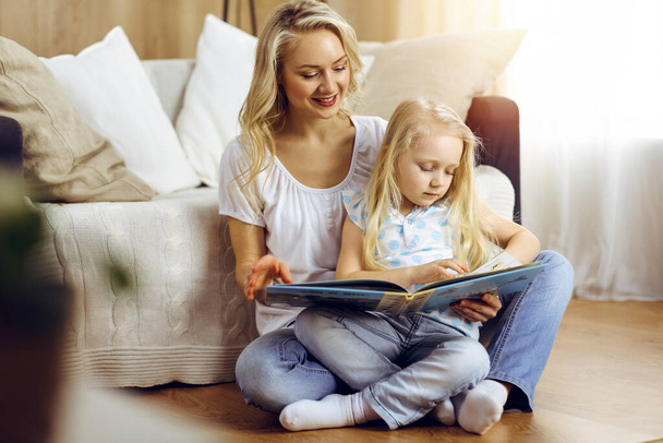 Onnellinen perhe. Blondi nuori äiti lukee kirjaa söpölle tyttärelleen istuessaan puulattialla aurinkoisessa huoneessa. Äitiyden käsite. - Valokuva, kuva