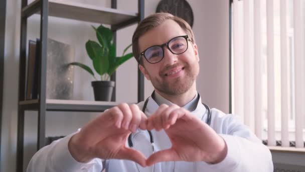 Portret van een lachende jonge huisarts in wit medisch uniform met een hart liefde handgebaar. Gelukkige jonge mannelijke dokter met steun. Het concept van de behandeling van mensen. - Video