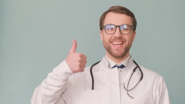 Un guapo doctor con uniforme médico blanco sobre fondo gris en el estudio muestra un gesto de mano. - Metraje, vídeo