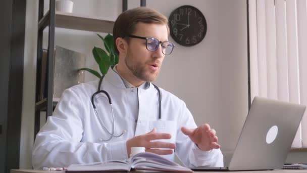 Ένας ιατρικός εργάτης κάθεται σε μια σύγχρονη κλινική, ντυμένος με λευκό παλτό γιατρού, κάνοντας βιντεοκλήση χρησιμοποιώντας ένα φορητό υπολογιστή, συμβουλεύοντας εξ αποστάσεως. - Πλάνα, βίντεο