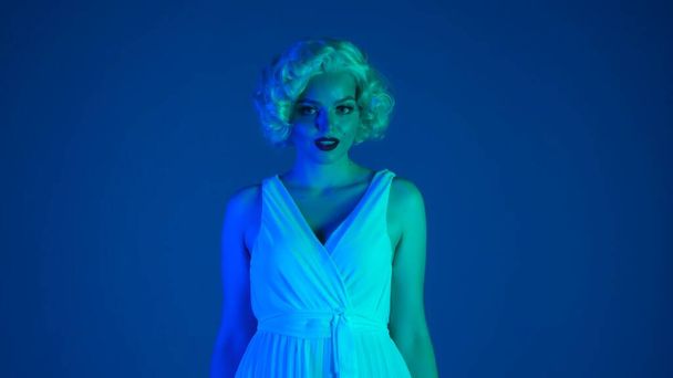 Portrait of a charming woman in the image of Marilyn Monroe close up. Woman with bright makeup in white dress and wig in studio in pink and green neon light - Фото, изображение