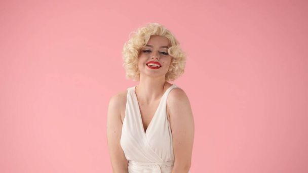 Portrait young woman in wig, white dress and with red lipstick on lips in studio on pink background. Woman looking like Marilyn Monroe in studio on pink background - Fotó, kép