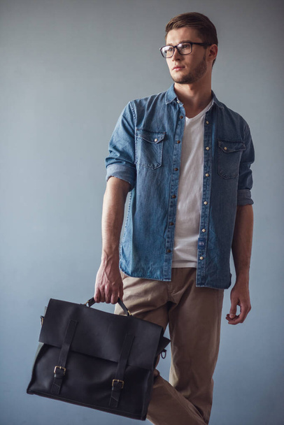 Portret van knappe jonge zakenman in casual kleding, brillen en met een stijlvolle tas, op de grijze achtergrond - Foto, afbeelding