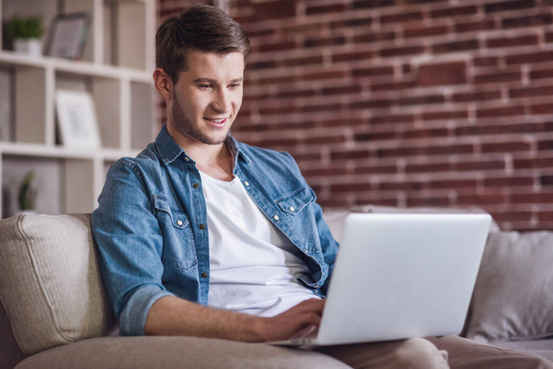 Przystojny facet w ubranie jest za pomocą laptopa i uśmiechając się siedząc na kanapie w domu - Zdjęcie, obraz