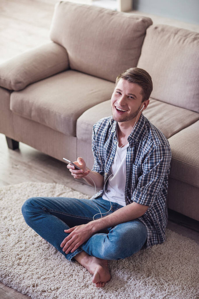 Όμορφος άντρας στα ακουστικά είναι χρησιμοποιώντας ένα έξυπνο τηλέφωνο και χαμογελώντας ενώ κάθεται στο πάτωμα στο σπίτι - Φωτογραφία, εικόνα