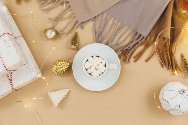 Cup of coffee, gift box and knitted sweaters, led lights on beige background. Hygge style, winter concept. Cozy home desk. New year and Christmas celebration mockup. Christmas still life - Zdjęcie, obraz