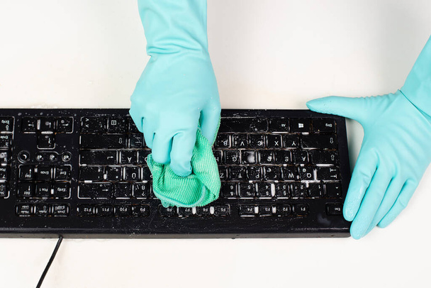 Ofisteki bilgisayar klavyesini plastik koruyucu eldivenle temizlemek, su ve sabunla silmek, tozlu ve kirli elektronik temizlik, ev işi.  - Fotoğraf, Görsel