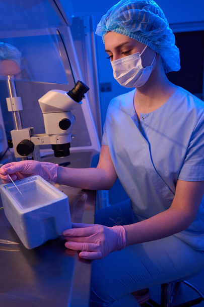 Naaraspuolinen alkiontutkija steriileissä käsineissä ja kasvonaamiossa, joka asettaa lasiaispillit kylmäsäilytyslaatikkoon - Valokuva, kuva