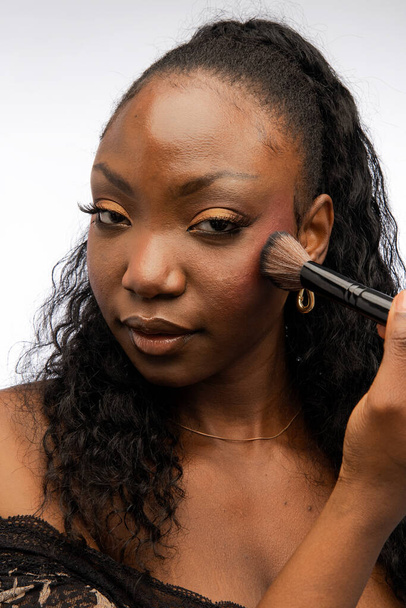 Νεαρή Αφρο-Αμερικανίδα γυναίκα που χρησιμοποιεί πινέλο καλλυντικών ενώ εφαρμόζει διακοσμητικό μακιγιάζ, κοιτάζοντας την κάμερα, απομονωμένη σε λευκό φόντο. Έννοια ομορφιάς και καλλυντικών - Φωτογραφία, εικόνα