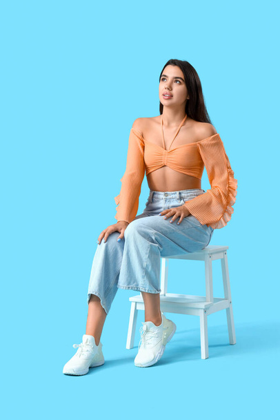 Beautiful young woman sitting on chair against blue background - Photo, Image